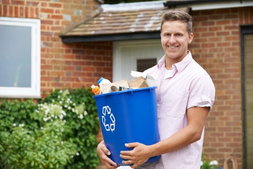 A young man with short, light brown hair and a friendly smile is standing outdoors in front of a red-brick house with white window frames and a brown tiled roof. He is wearing a light pink, short-sleeved shirt and holding a large blue recycling bin with a white recycling symbol on the side, filled with various pieces of waste including cardboard, plastic bottles, and paper. The bin is positioned in the foreground, while the background features green bushes with white flowers and a paved driveway, suggesting a residential setting in Stratford or nearby within the UK. The scene captures a clear, bright day with natural light illuminating the individual's face and the surroundings, reinforcing the context of rubbish collection or house clearance services offered by Stratford House Clearance.