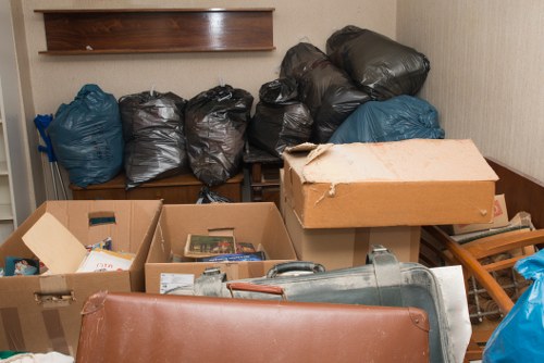 The image shows a cluttered indoor space, possibly a storage or utility room, with a variety of rubbish bags and boxes. In the background, there are six large black and blue garbage bags stacked against a wooden panel or shelf, with some bags appearing full and sealed. In the foreground, several open cardboard boxes contain miscellaneous household items, including books, papers, and packaging materials. A large, transparent plastic container with a handle is partially visible among the boxes. To the right, there is a wooden drying rack or clothes airer leaning against the wall, and a rolled-up piece of cardboard or padding is resting on top of some items. The lighting appears natural or diffused, illuminating the cluttered environment in a neutral tone. This scene exemplifies equipment and materials typical of rubbish clearance carried out by Stratford House Clearance in the Stratford area, demonstrating their capacity to manage varied household waste and debris for efficient rubbish removal services in London.