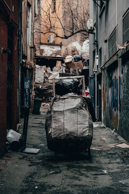 Two large black plastic garbage bags filled with waste material are placed on the pavement beside a black metal fence. The bags appear slightly crumpled and are positioned one leaning against the othe