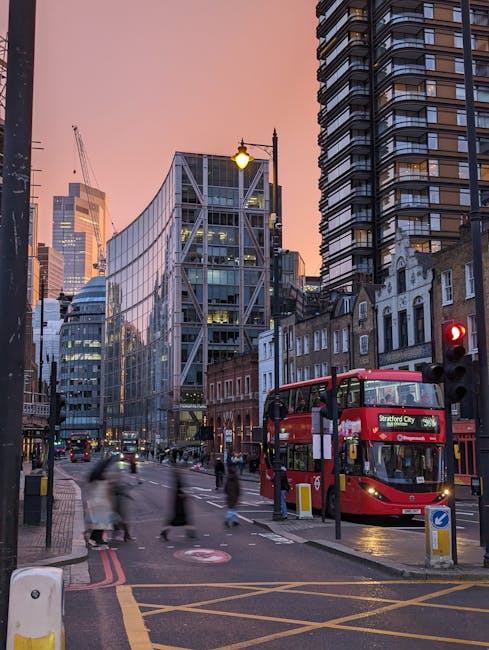 A city street scene at dusk featuring a red double-decker bus labeled 'Stratford City' parked at a bus stop. In the foreground, a traffic light displays a red signal, and pedestrians are crossing the 