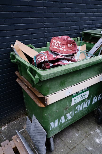 A green wheeled rubbish bin filled with red and black bags of building or garden waste positioned outdoors on a paved surface against a dark navy brick wall. The bin contains bags labeled with product