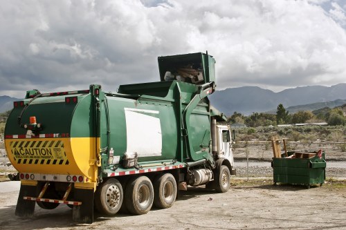 A large green waste collection truck with a yellow caution stripe and black hazard markings on the rear is parked on a gravel or dirt surface, with its rear loading compartment open and positioned to collect rubbish. To the right of the truck, there is a green wheeled waste bin filled with assorted debris, likely part of a rubbish removal service. The background features a landscape of hills or mountains under a partly cloudy sky, suggesting a semi-rural or outskirts environment. The setting appears to be an outdoor waste collection or disposal site, consistent with professional rubbish clearance activities often undertaken by companies such as Stratford House Clearance in Stratford, serving local waste removal needs.