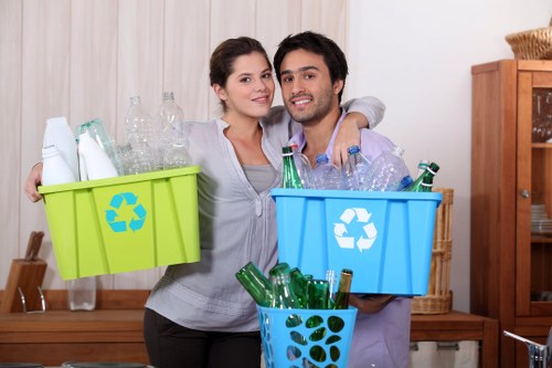 A young couple stands indoors, each holding a plastic recycling bin filled with used glass bottles; the woman’s bin is bright green while the man’s is blue, both featuring the universal recycling symbol on the side. The woman has light brown hair pulled back and wears a light grey shirt, smiling softly. The man has dark, slightly wavy hair, and facial hair, dressed in a lavender shirt, smiling broadly. They are positioned close together, with one arm around each other’s shoulders, facing the camera. Behind them, a wooden sideboard and a shelf with various items are visible, set against a cream-colored wall, suggesting a domestic kitchen or living area. The bins are filled with clear and green glass bottles, some with labels visible, indicating they are ready for recycling. The lighting in the room is bright and even, highlighting the textures of the bottles and the smooth surfaces of the bins. This image visualizes the act of glass bottle recycling, a service often associated with waste management companies like Stratford House Clearance, operating to promote environmentally responsible rubbish disposal in Stratford and surrounding areas.