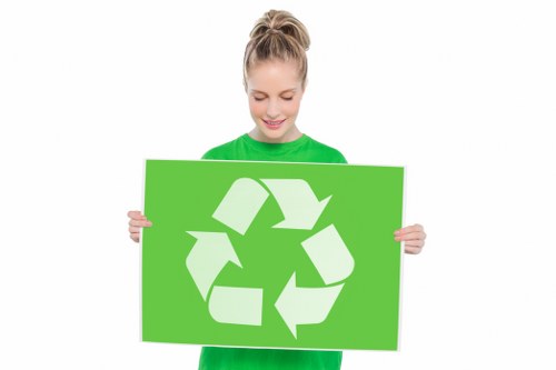 A young woman with blonde hair tied back in a ponytail, wearing a bright green long-sleeved top, holding a large lime-green sign featuring a white recycling symbol composed of three arrows forming a triangle. She is smiling gently and looking down at the sign, standing against a plain white background. The image emphasizes environmental awareness and recycling, which is relevant to waste management and rubbish removal services offered by Stratford House Clearance near Stratford, with a focus on sustainability and responsible disposal practices.